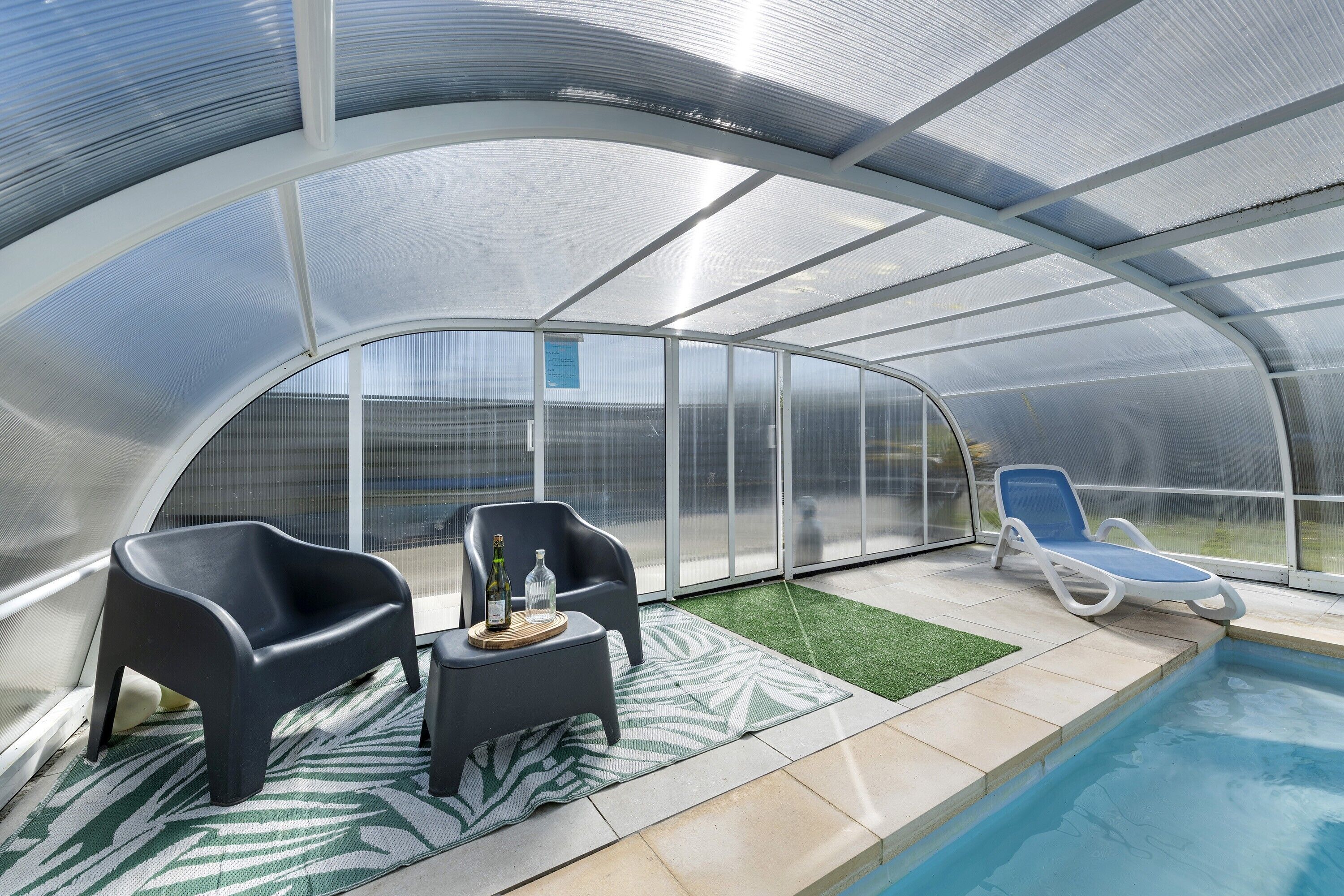 A covered outdoor area with a pool, two lounge chairs, and a small table with a bottle and glass.