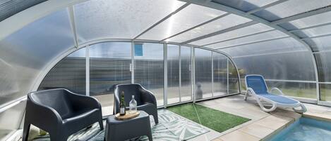 A covered outdoor area with a pool, two lounge chairs, and a small table with a bottle and glass.