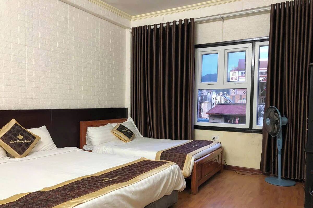 Habitación de hotel con dos camas, una ventana con cortinas, un ventilador y vistas a edificios.