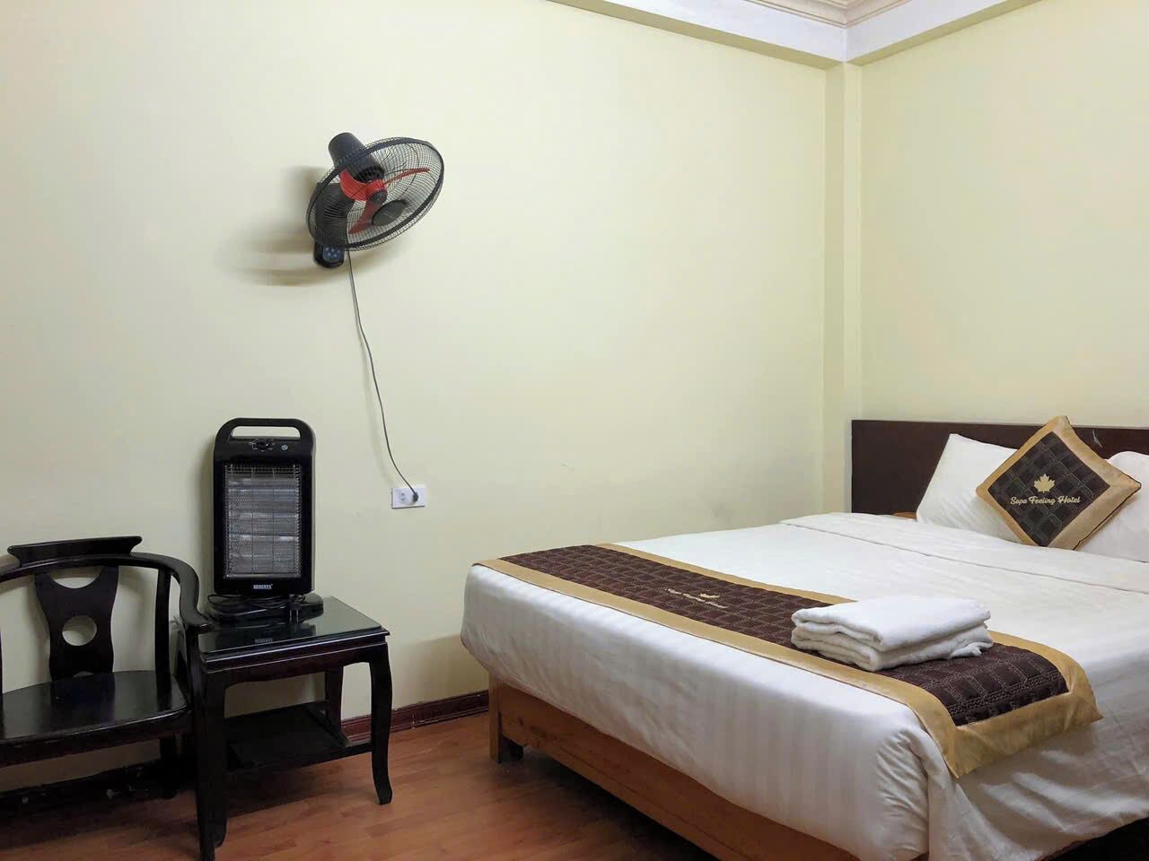 A hotel room with a bed, a desk with a heater, and a wall-mounted fan.
