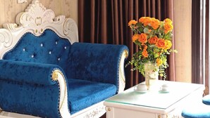 Une pièce avec un canapé en velours bleu, une table blanche ornée d’un vase de fleurs oranges et un lit recouvert de draps blancs.