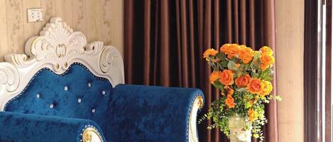 Una habitación con un sofá de terciopelo azul, una mesa blanca con un jarrón de flores naranjas y una cama con sábanas blancas.