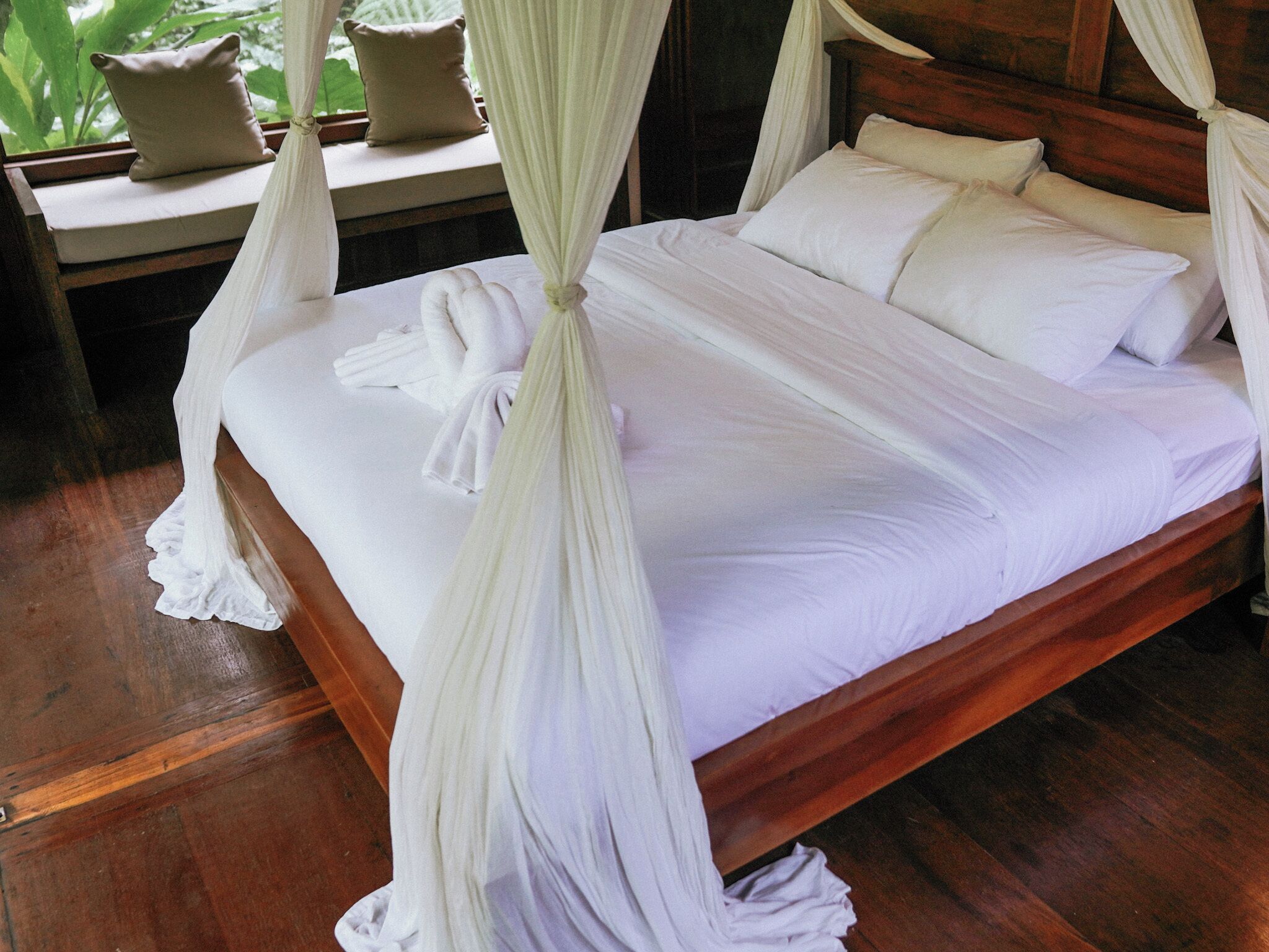 Un lit avec du linge de lit blanc et un baldaquin, une tête de lit en bois et une fenêtre donnant sur un paysage verdoyant.