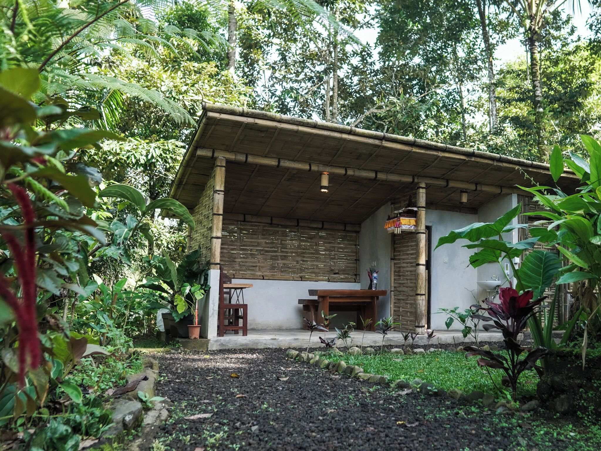 A small, rustic building with a bamboo roof and a concrete foundation, surrounded by lush greenery.