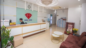 A hotel reception area with a modern design, featuring a reception desk, a chandelier, and a seating area with a sofa and a small round table. - The Sóng - Ruby Star Hotel Apartment (Ho Chi Minh City)