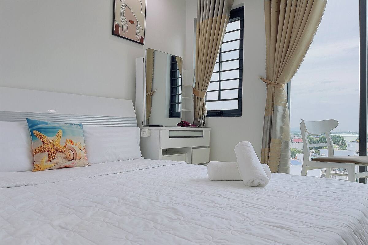 Habitación de hotel con cama, escritorio, silla, ventana con cortinas y vista a la ciudad.