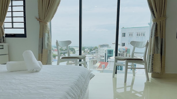 A hotel room with a large bed, two white chairs, and a view of the cityscape through large windows. - The Sóng - Ruby Star Hotel Apartment (Ho Chi Minh City)