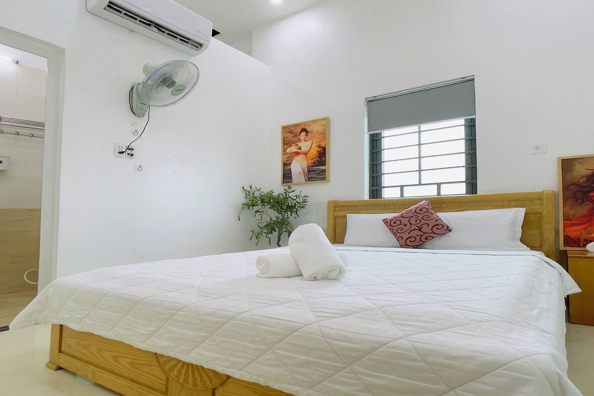 Una cama bien tendida con colcha blanca, un ventilador de pared, una ventana con persianas y un cuadro en la pared.