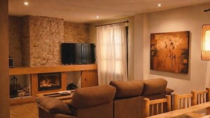 A cozy living room with a stone wall, a flat-screen TV, a fireplace, a large sofa, and a dining table. - Chalet in Rivero de Posadas with Pool (Rivero de Posadas)