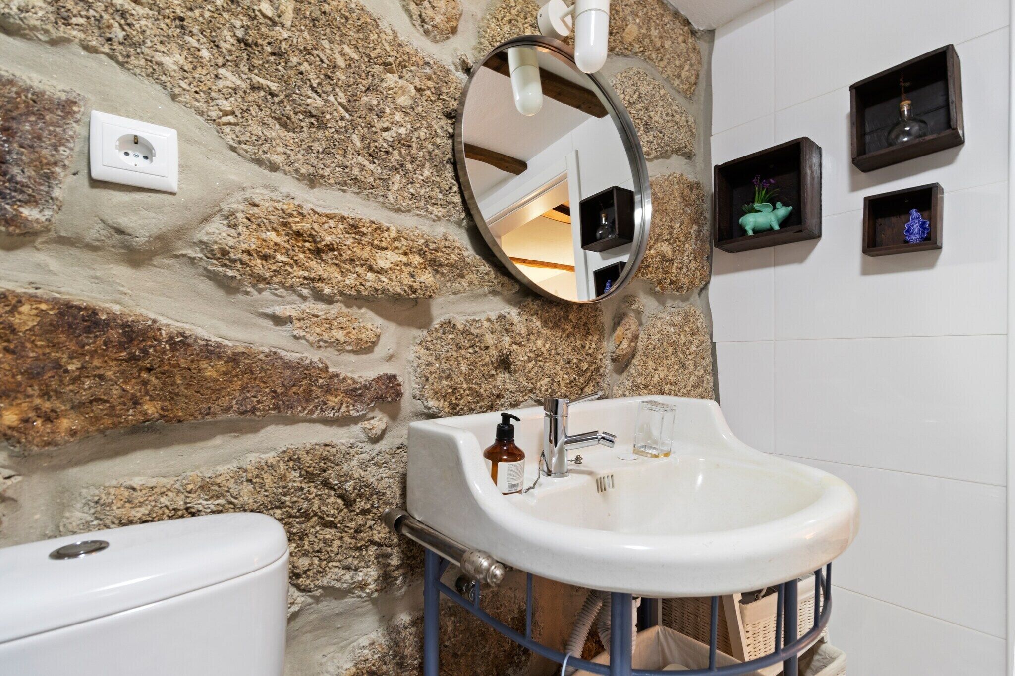 A bathroom with a stone wall, a white sink with a mirror above it, and three small shelves with decorative items.