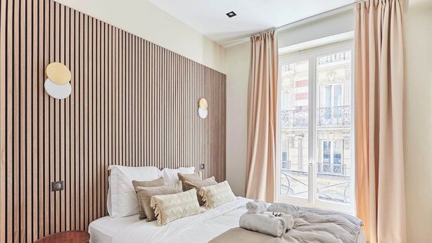 Ett modernt hotellrum med en stor säng, en väggpanel i trä, ett litet sidobord och ett fönster med gardiner.