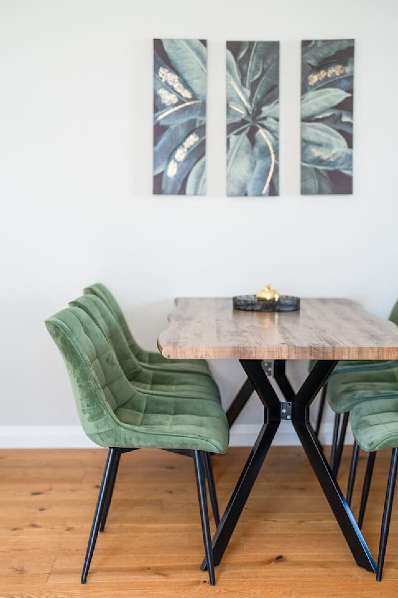A dining table with a wooden top and black metal legs, flanked by two green upholstered chairs. Above the table, there are two framed botanical prints on the wall.