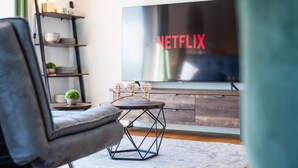 A modern living room with a large flat-screen TV displaying the Netflix logo, a comfortable gray sofa, a small round table with decorative items, and a wooden cabinet. - 90m2 Loft I Kaffee I Terrasse I E-Parkplatz (Rosenheim)