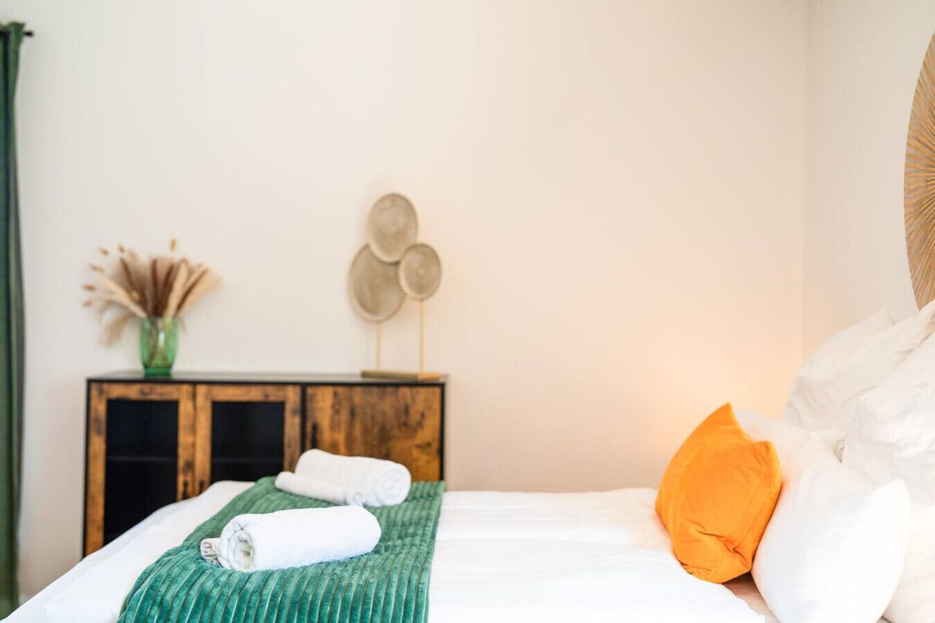 A neatly made bed with a green striped blanket, two rolled white towels, and an orange pillow.
