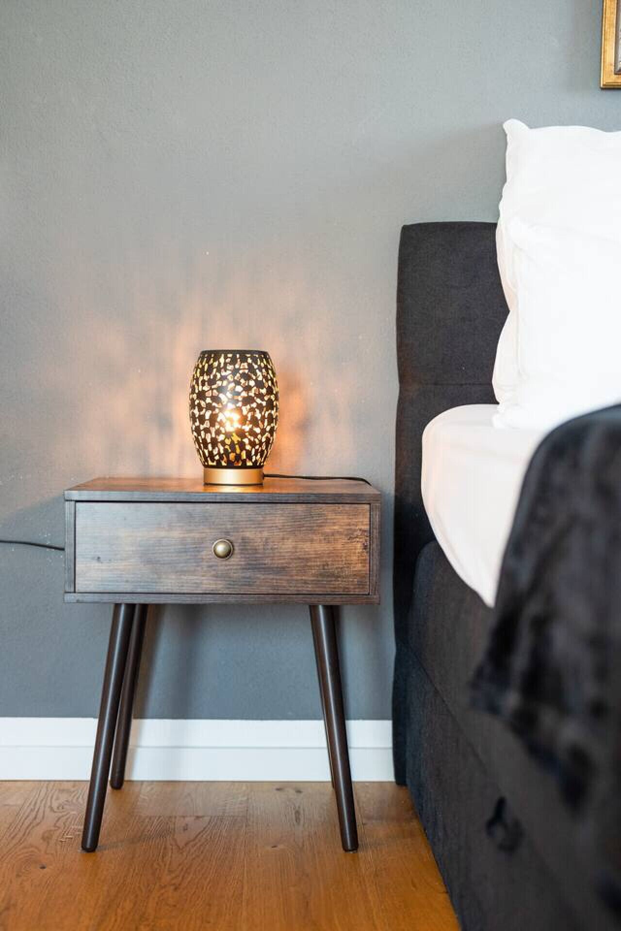 Une table de chevet avec une lampe à texture, un tiroir et une table de nuit de couleur sombre, le tout placé contre un mur gris.