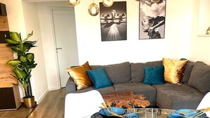 A modern dining area with a glass table set for six, surrounded by a grey sofa with blue and gold cushions. - YAYstay - 58m2 I balkony I 56` Smart TV I train (Mühldorf am Inn)