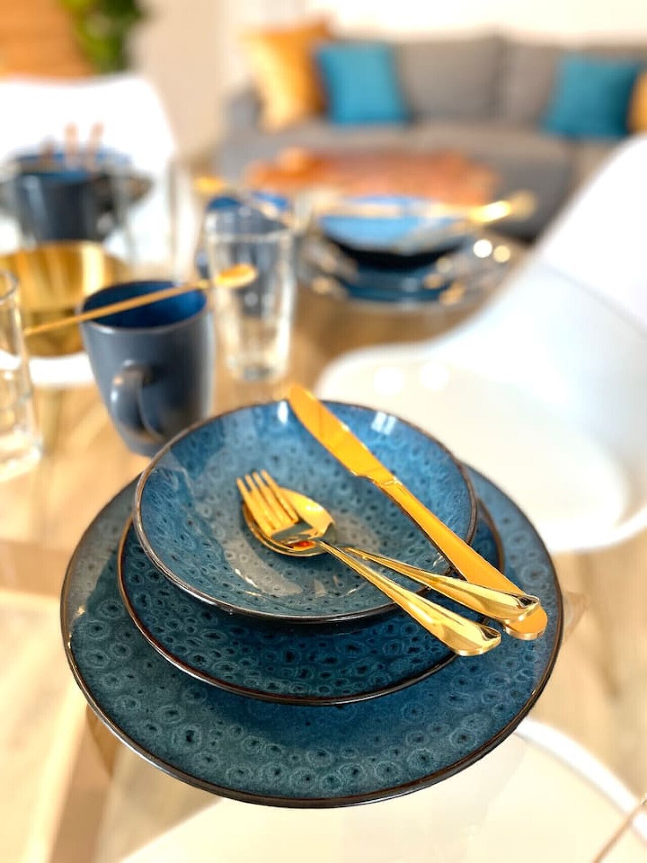 Ein Esstisch gedeckt mit blauen Tellern, goldbesteck und klaren Gläsern.