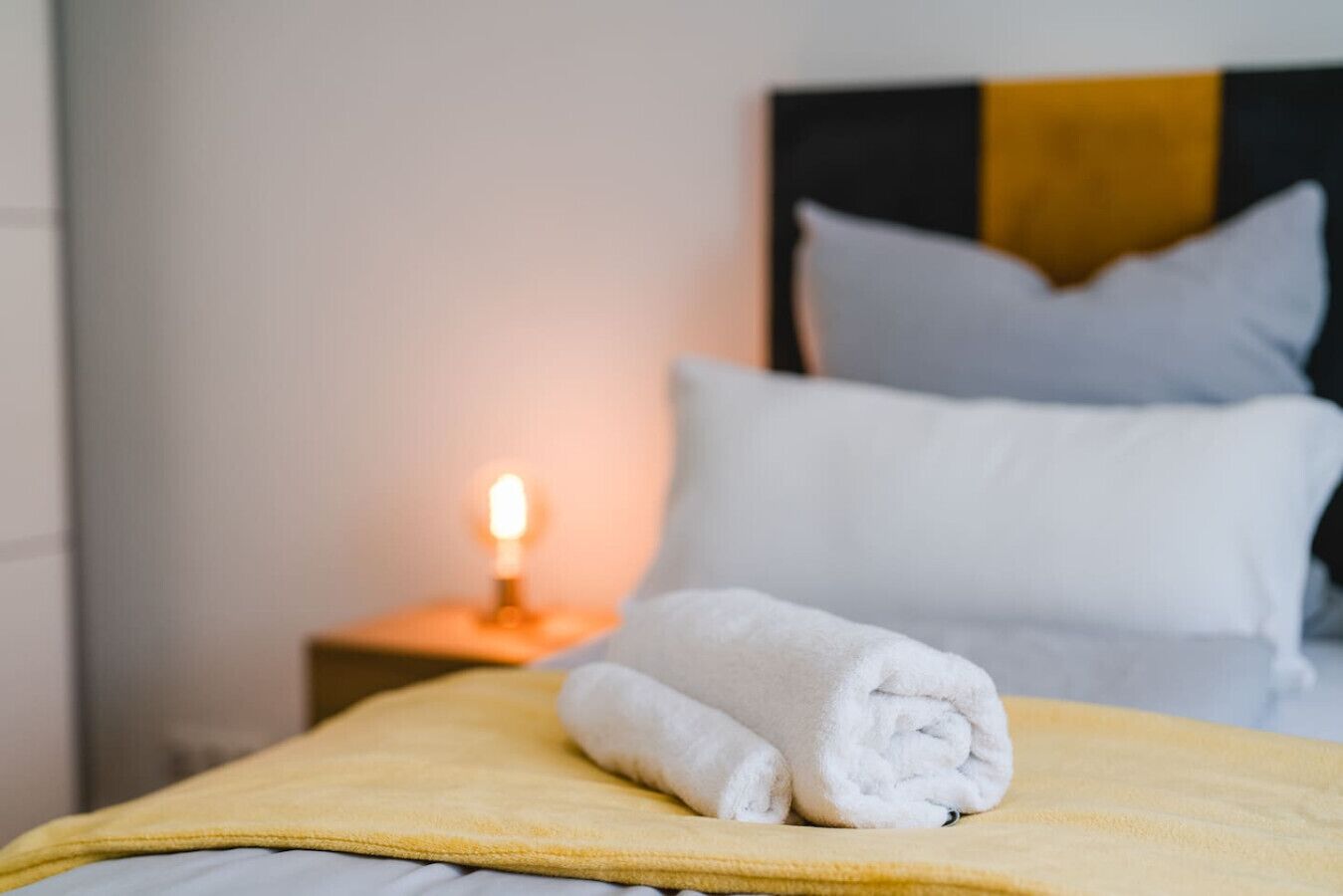 Un lit bien fait, recouvert d’une couverture jaune et agrémenté de serviettes blanches pliées, une table de chevet avec une bougie allumée et une tête de lit ornée d’oreillers.