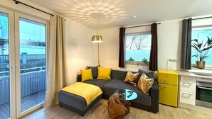 Una sala de estar moderna con sofá, reposapiés y mesa de centro, una pequeña cocina y un balcón con vistas.