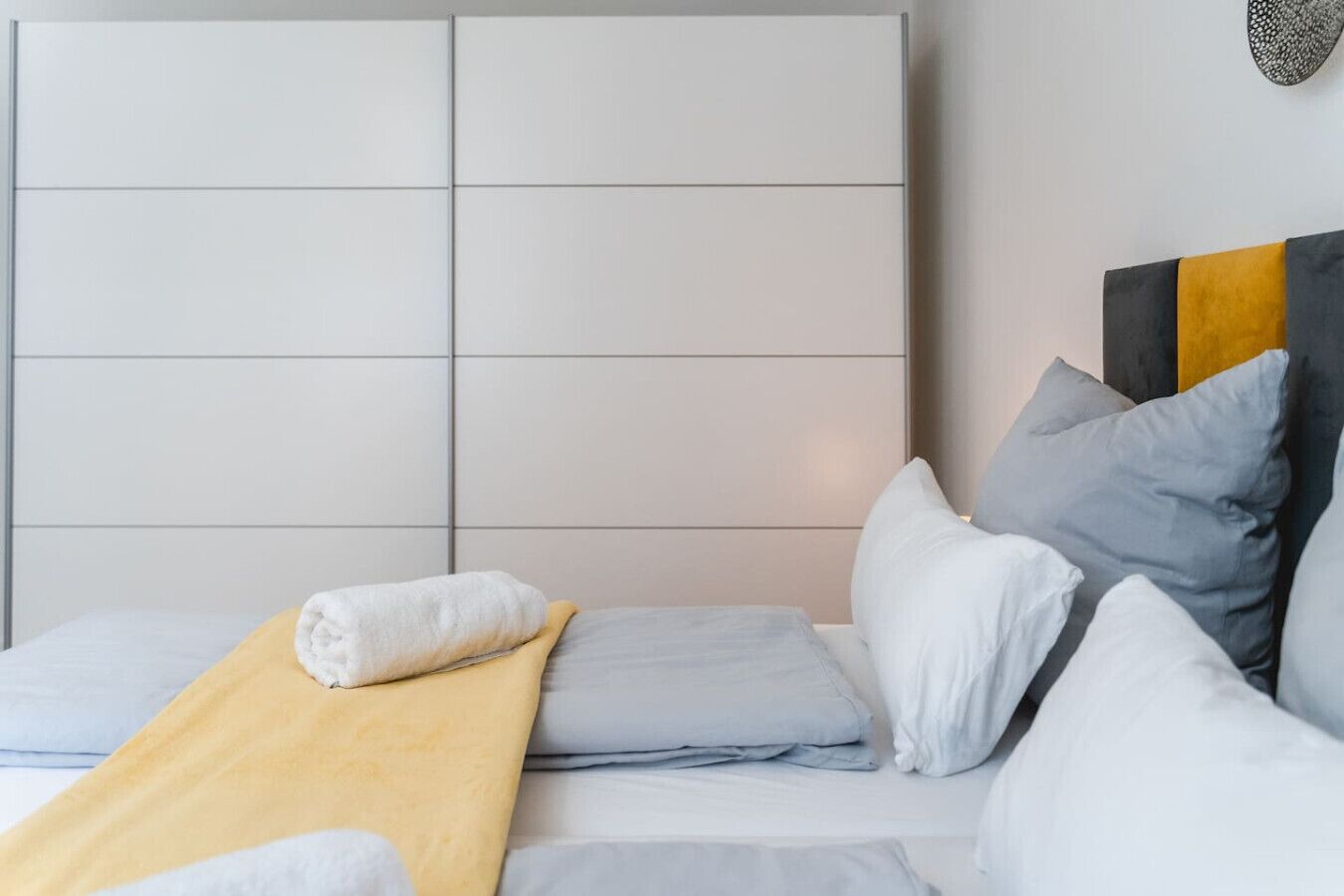 A modern bedroom with a large, padded headboard, a neatly made bed with a yellow throw blanket, and a variety of pillows in different shades.