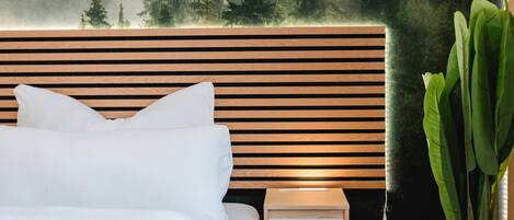 A modern bedroom with a wooden headboard, white bedding, a bedside table, and a large green plant.