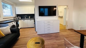 Un appartement moderne comprenant une cuisine, un coin repas et un salon. Il est équipé d’un canapé, d’une table basse et d’un téléviseur fixé au mur.