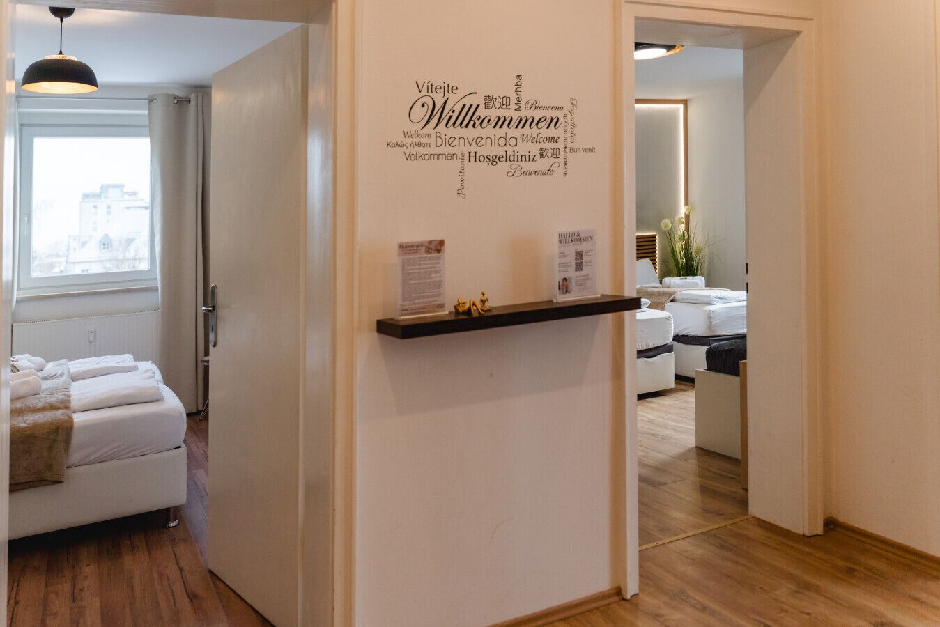 Une chambre d’hôtel comprenant un lit, une table de chevet, une étagère fixée au mur avec une pancarte, et une fenêtre avec des rideaux.