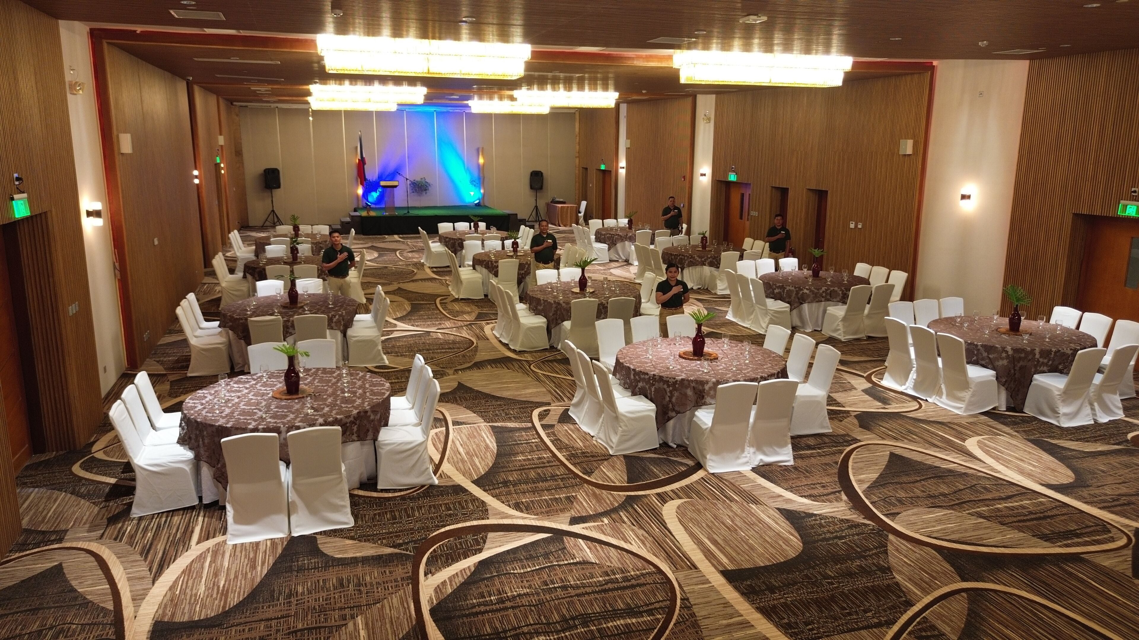 A banquet hall with round tables covered in tablecloths, white chairs, and a stage with blue lighting in the background.