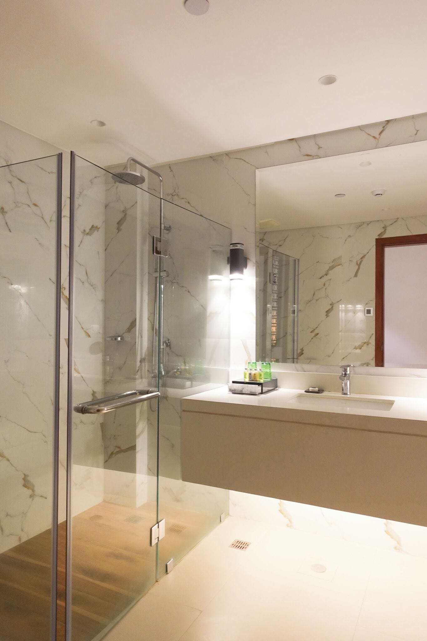 A modern bathroom with a glass-enclosed shower, a marble-tiled wall, a large mirror, and a countertop with toiletries.