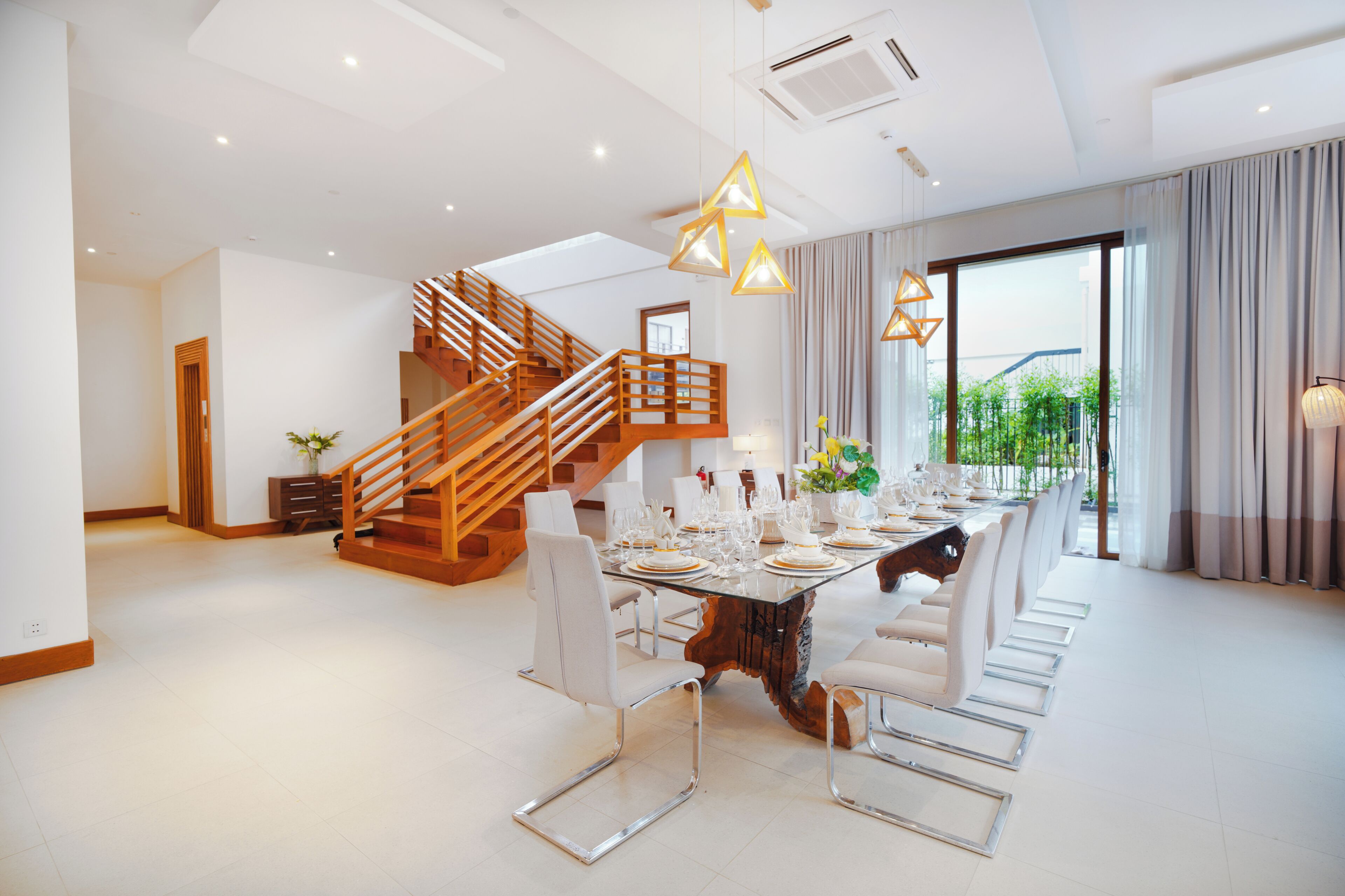 A spacious dining area with a long table set for a meal, a wooden staircase, and large windows with curtains.