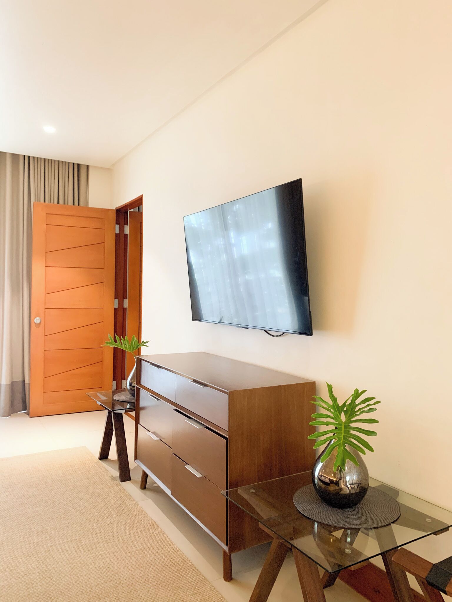 A modern living room with a flat-screen TV mounted on the wall, a wooden cabinet, a glass coffee table, and a potted plant.