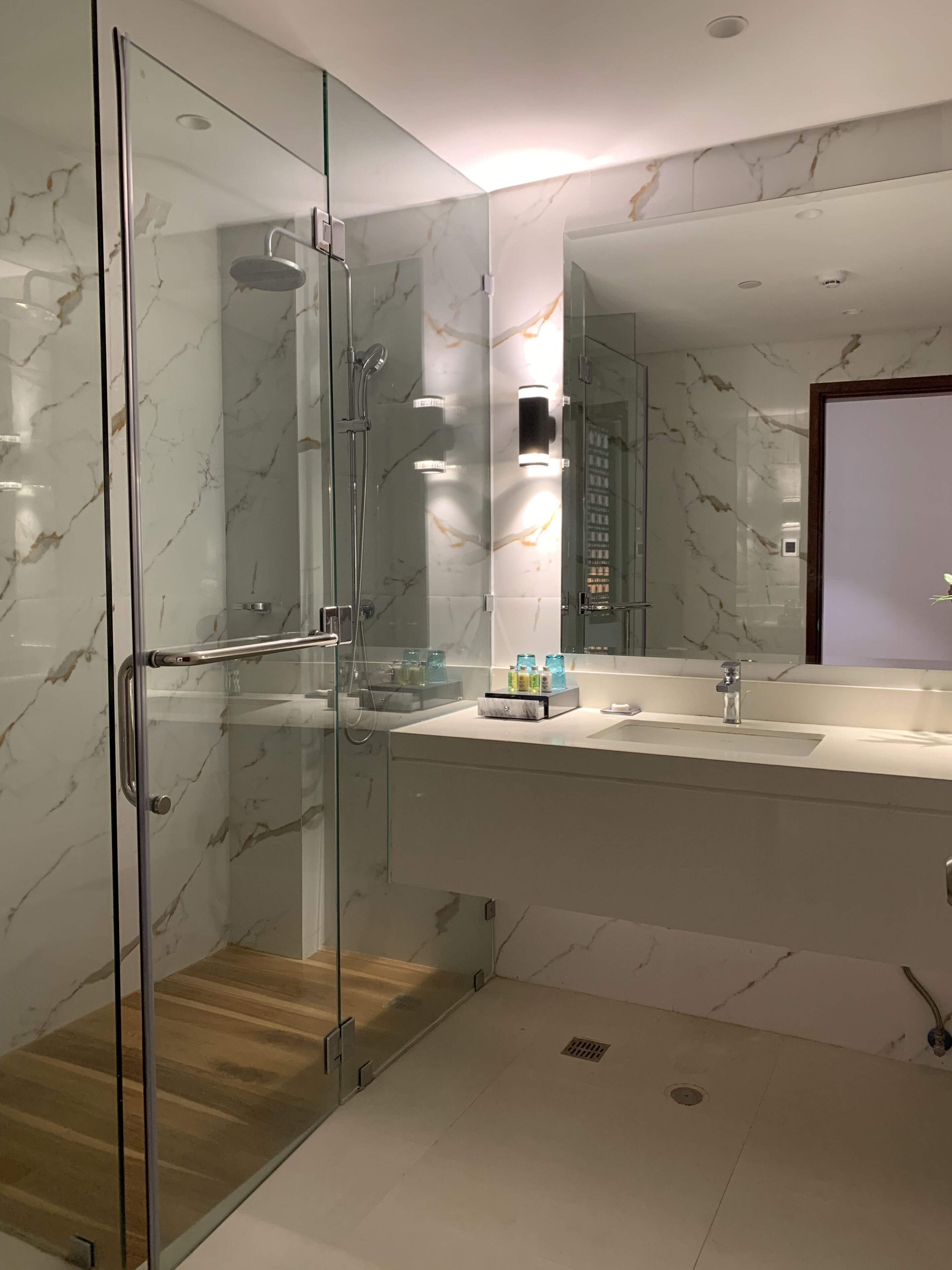A modern bathroom with a glass-enclosed shower, a marble-tiled wall, a large mirror, and a wooden floor.