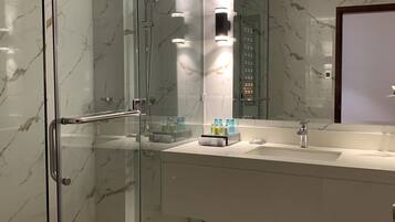 A modern bathroom with a glass-enclosed shower, a marble-tiled wall, a large mirror, and a wooden floor.
