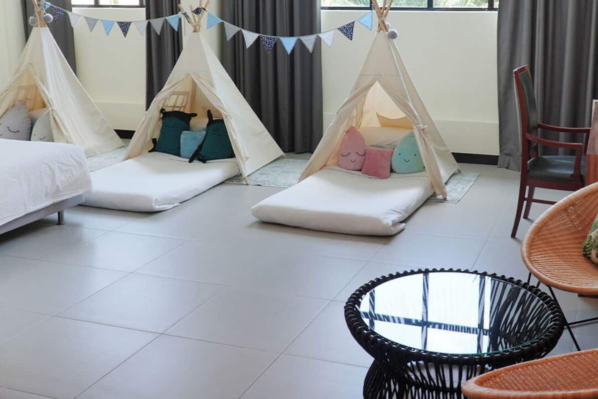 A room with three sleeping areas, each with a tent-like structure and pillows. There is a round glass table and wicker chairs.