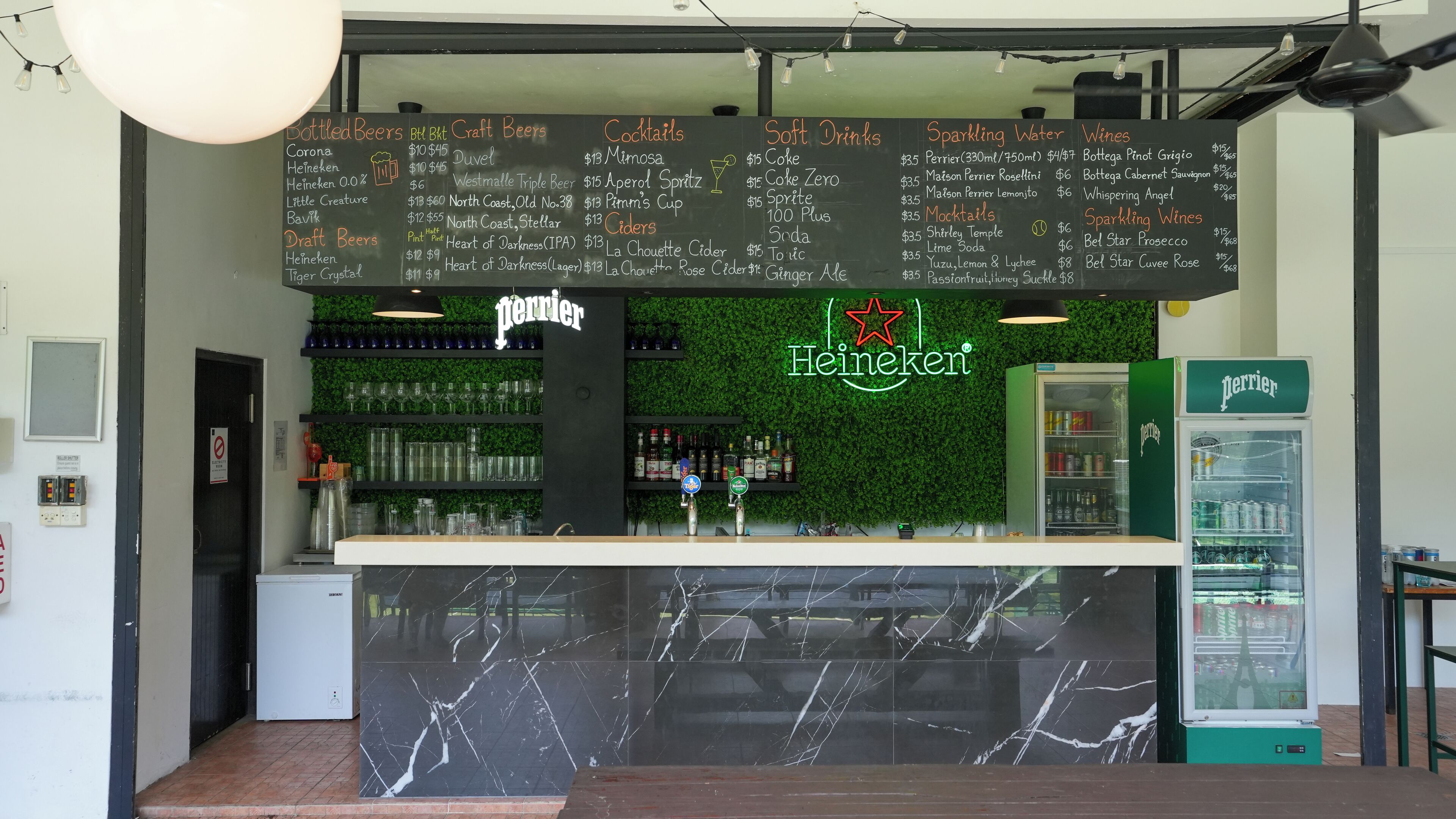 Zona de bar con una pizarra que muestra un menú de bebidas varias y un refrigerador de Perrier.
