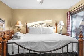 Ein ordentlich bezogenes Bett mit weißen Leinentüchern, einem Holzkopfende und einem Metallgestell. Es gibt zwei Nachttischlampen und ein Fenster mit einem karierten Vorhang.