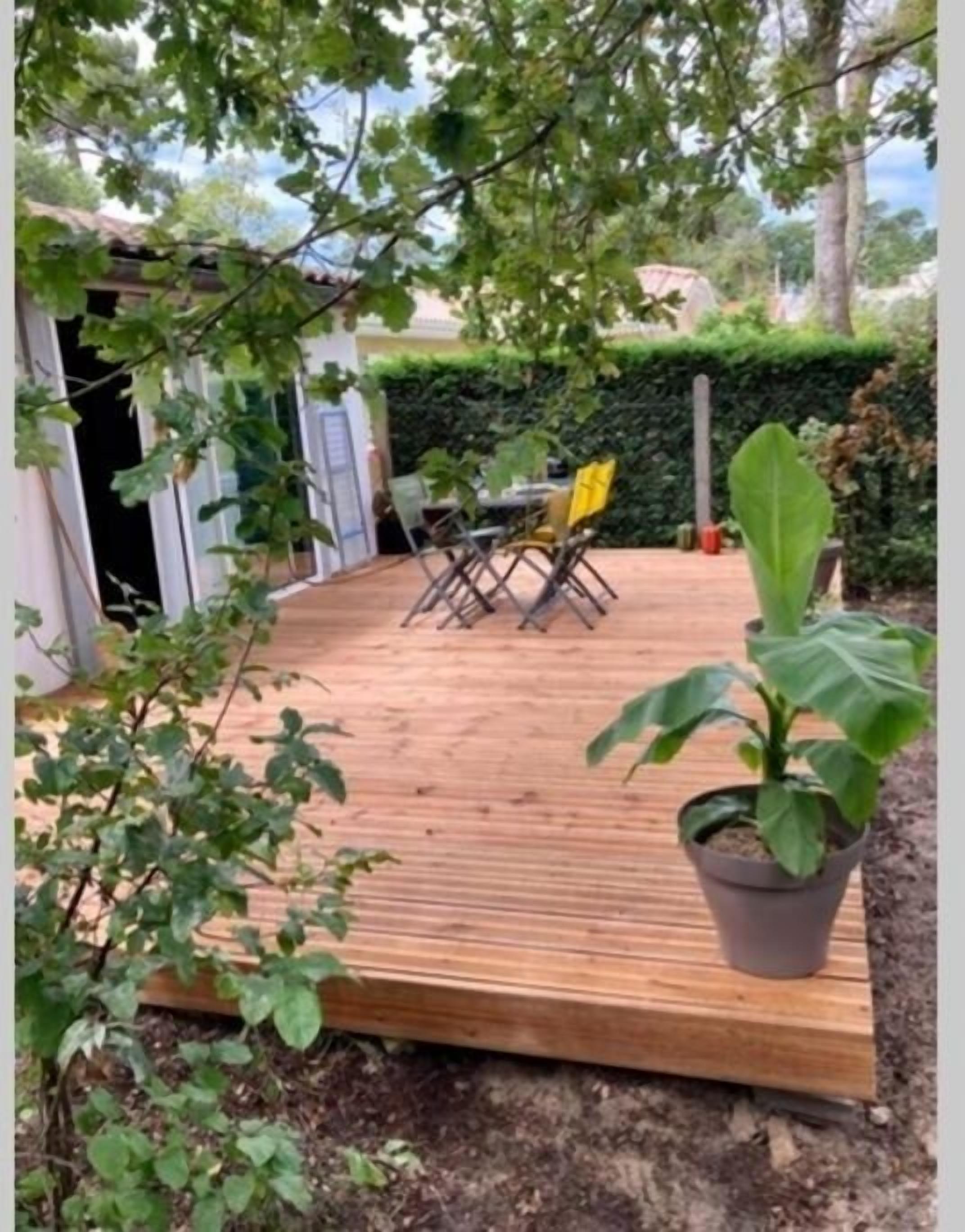 Una terraza de madera con una mesa y sillas, rodeada de follaje y un seto.