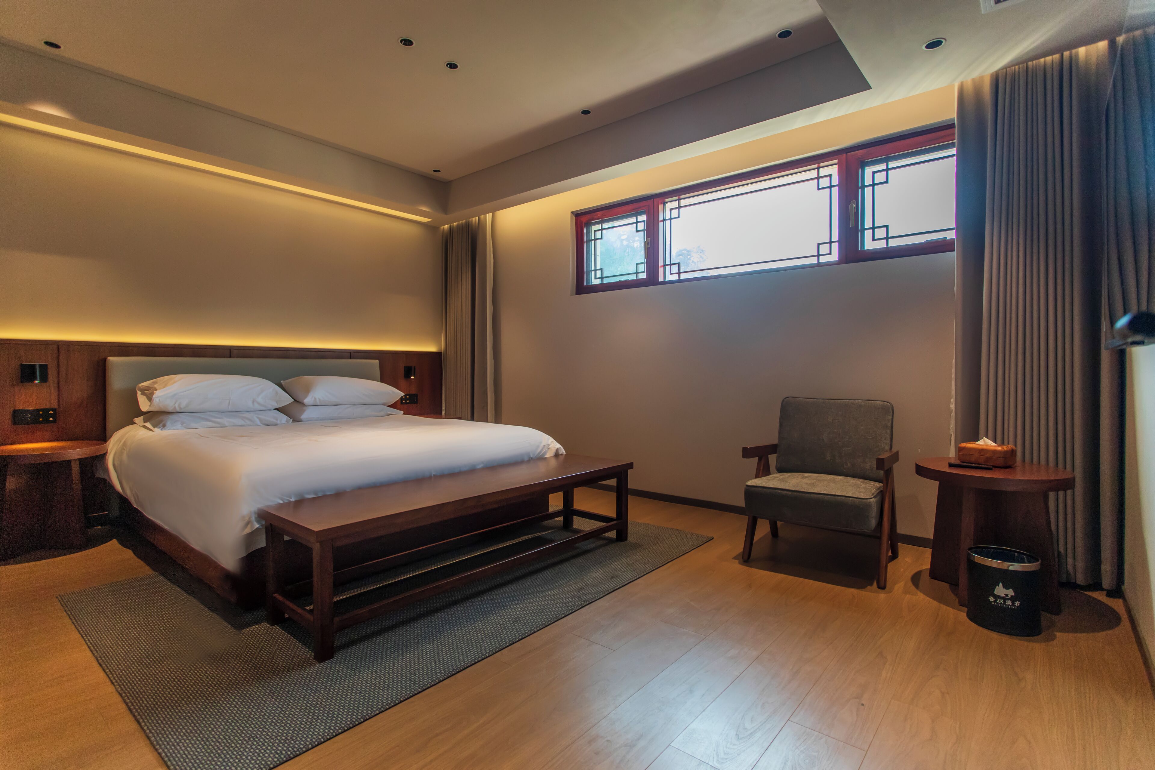 Una habitación de hotel moderna con una cama grande, una silla, una mesita y un ventanal con cortinas.