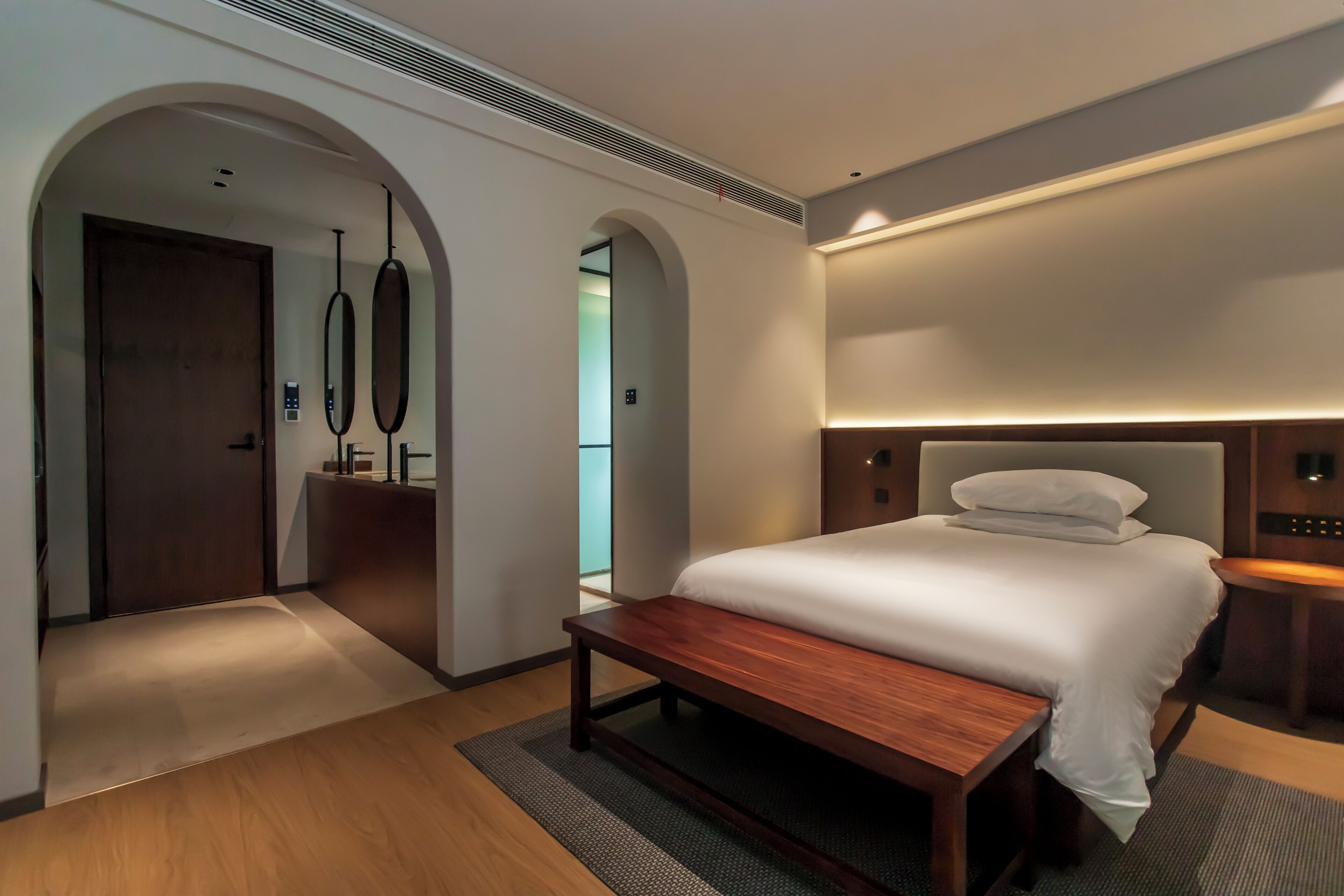 Habitació d'hotel moderna amb un llit gran, una banca de fusta i un escriptori. Hi ha portes arqueades i una làmpada de paret.
