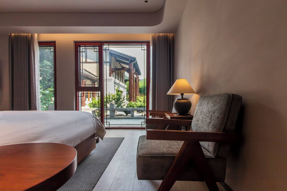 A hotel room with a bed, a chair, a table, and a lamp. There is a view of a building and greenery outside the window.