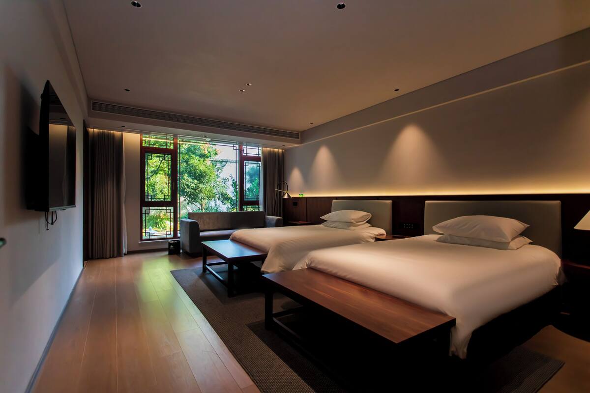 A modern hotel room with two beds, a flat-screen TV, a bench, and a large window offering a view of greenery.