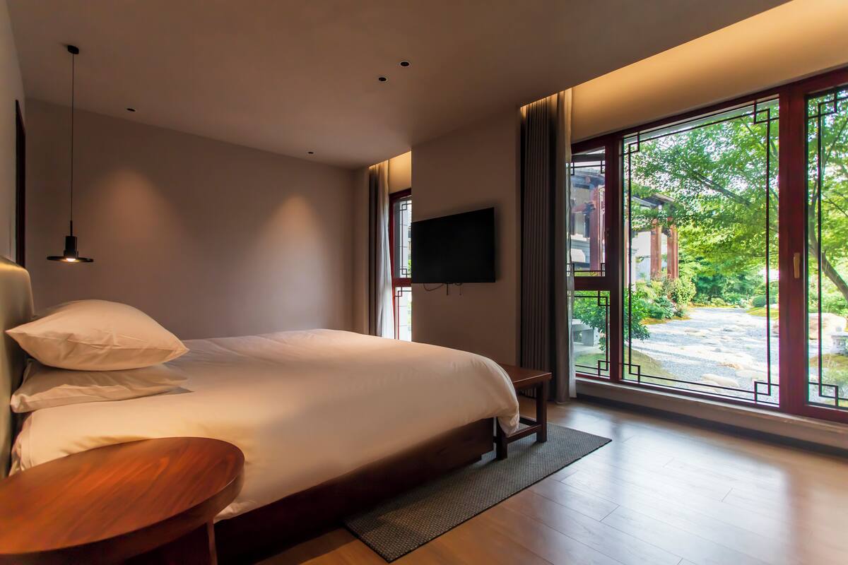 A modern bedroom with a large bed, a TV mounted on the wall, and a view of a garden through large windows.