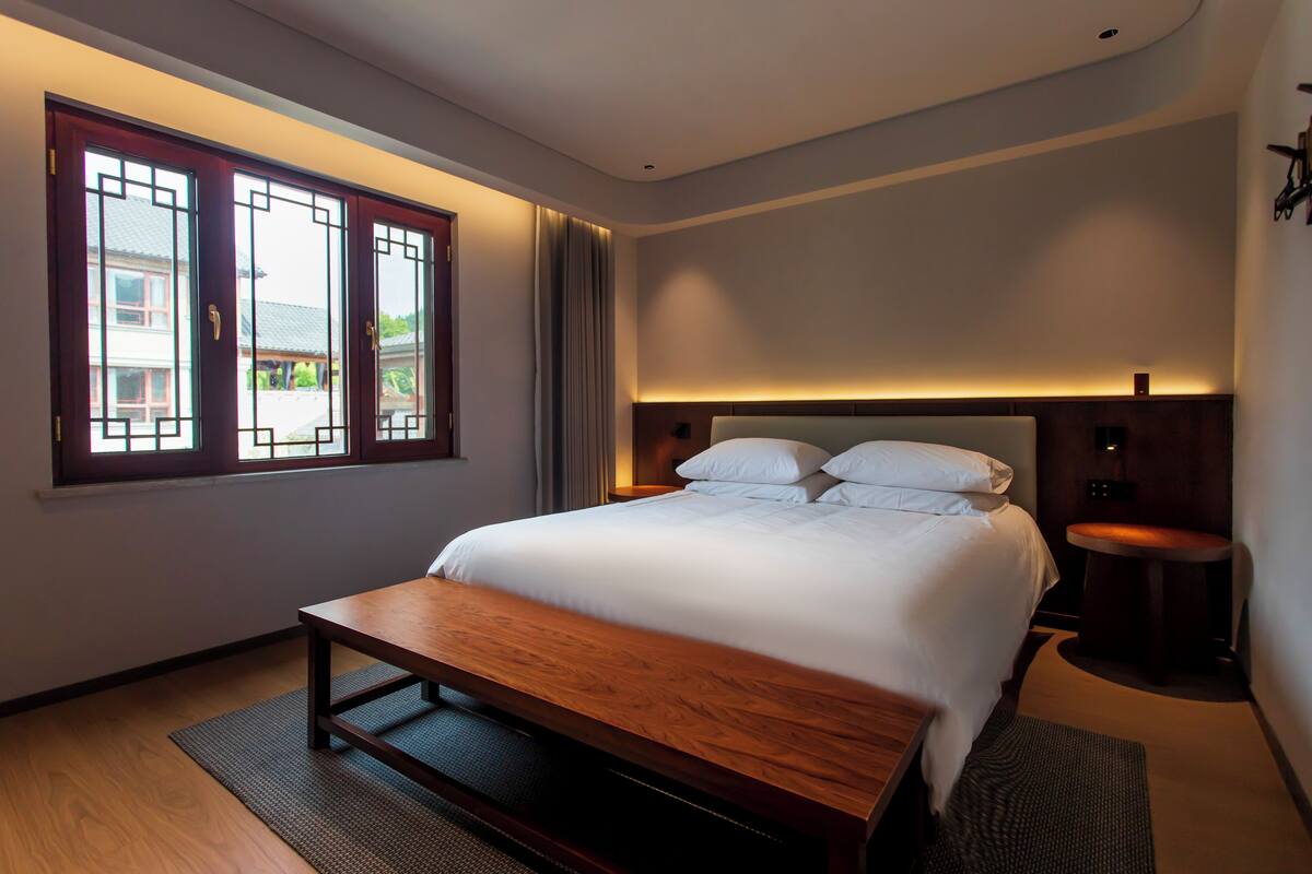 A modern hotel room with a large bed, a wooden bench, a small table, and a view of the outside through a window with decorative glass panels.