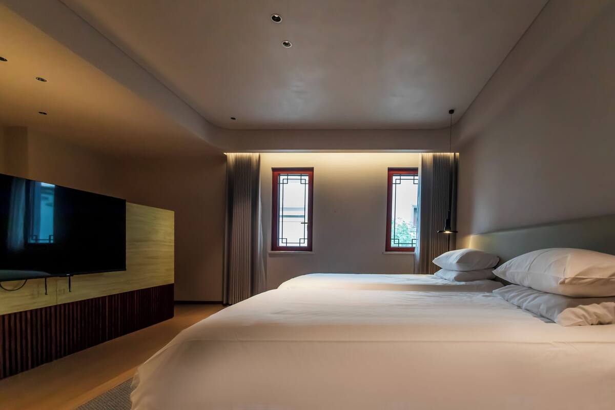 A modern hotel room with a large bed, a flat-screen TV mounted on a wooden panel, and two windows with curtains.