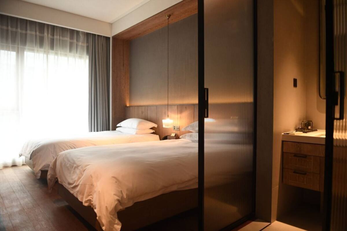 A hotel room with two beds, a wooden headboard, a bedside table with a lamp, and a view of the outside through sheer curtains.