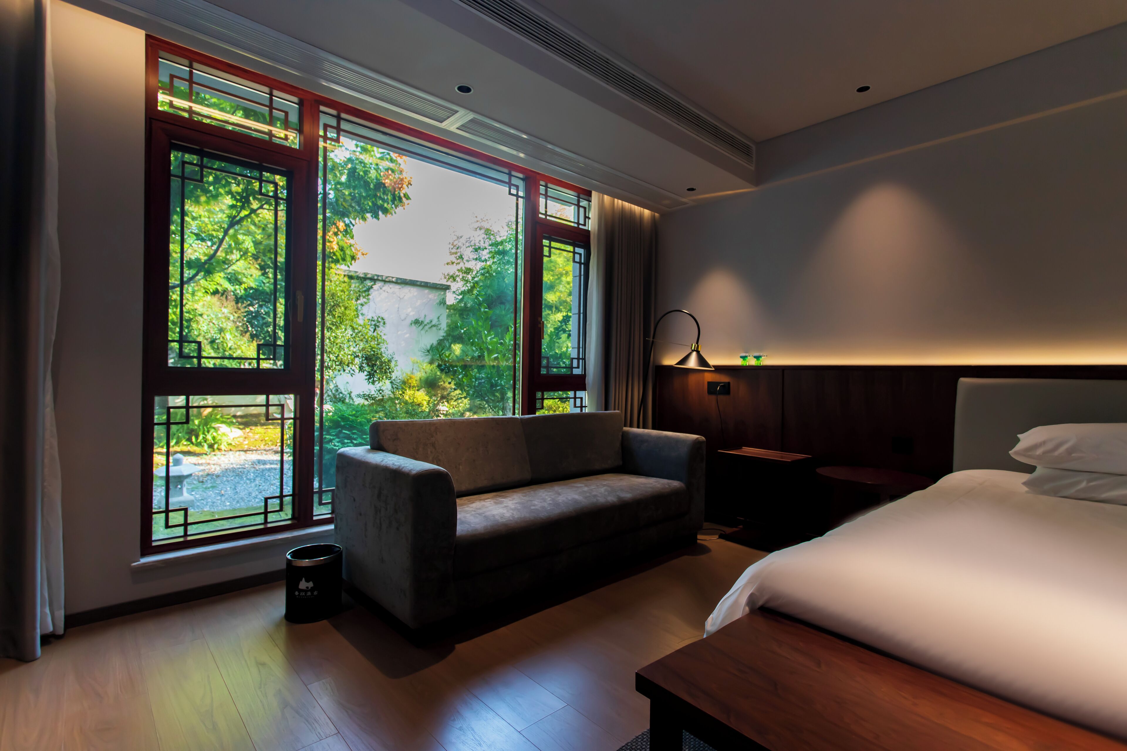 Ein modernes Hotelzimmer mit einem großen Bett, einer Couch, einem kleinen Tisch und einem Nachttisch. Große Fenster bieten Blick auf die grüne Umgebung.