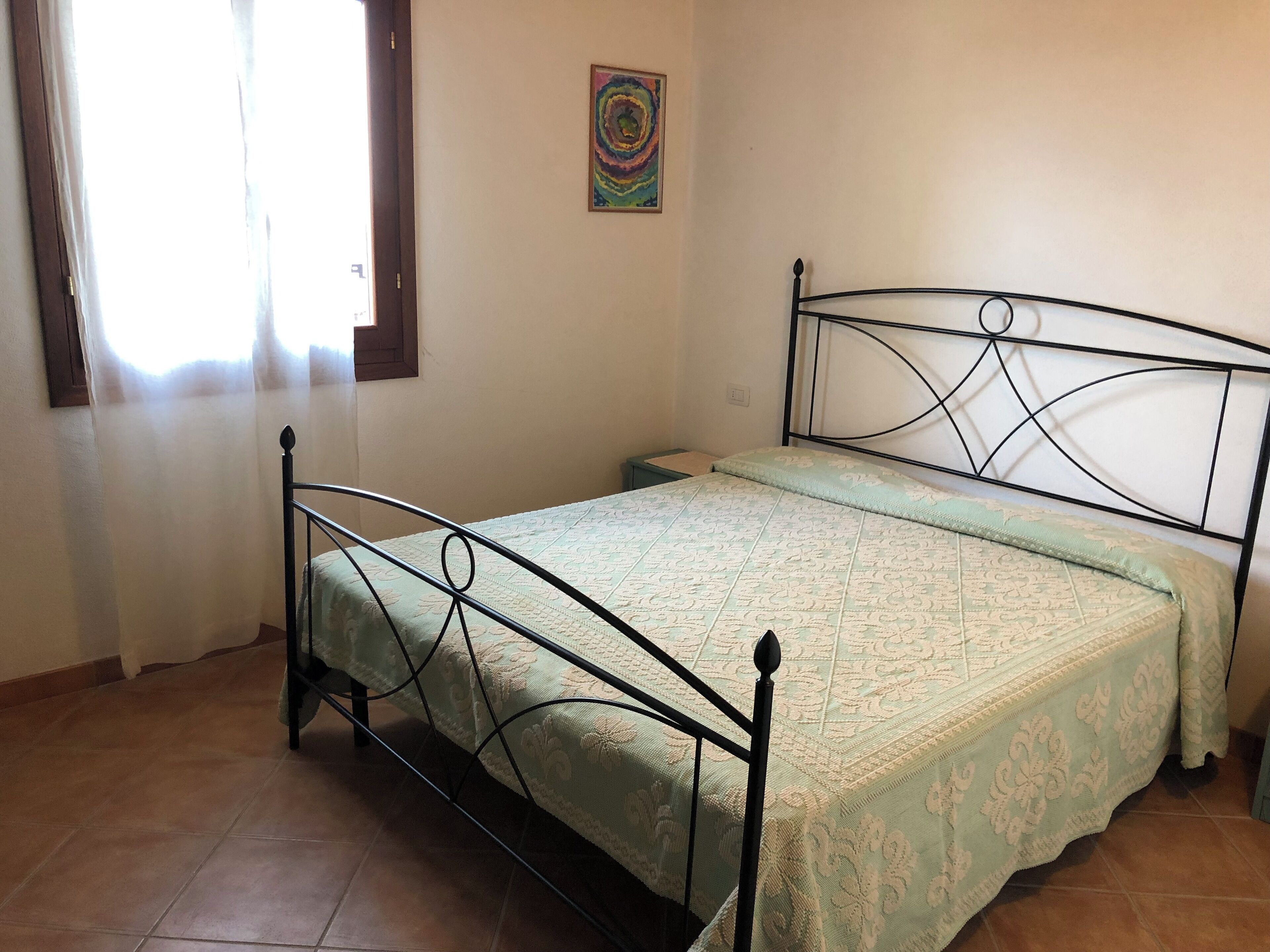 Ein Zimmer mit einem Einzelbett, einem Metallgestell, einer gemusterten Bettdecke, einem kleinen Nachttisch und einem Wandbild.