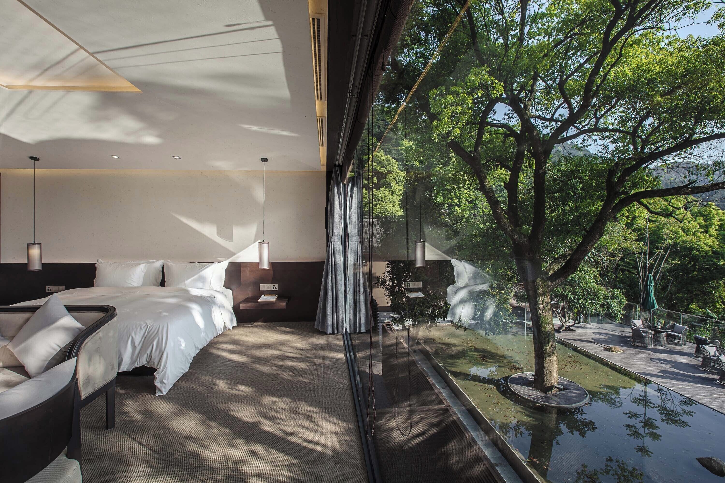 A modern hotel room with a large bed, a sitting area, and a view of a garden through a glass wall.