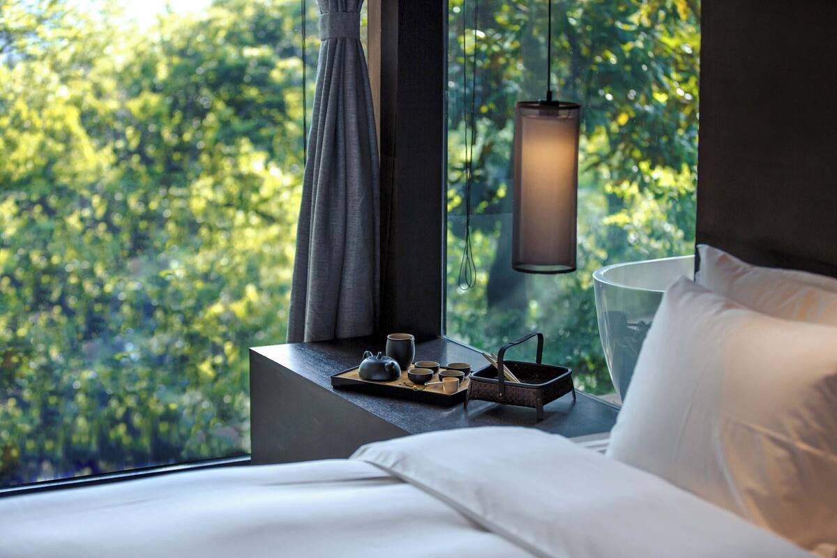 A neatly made bed with white linens, a bedside table with a tray holding a teapot, cups, and a small black container, and a hanging lamp.
