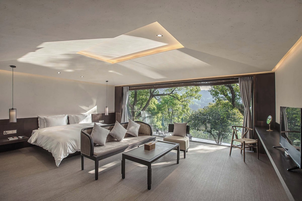 A modern hotel room with a large bed, a sofa, a coffee table, and a TV. The room has a large window with a view of trees and mountains.